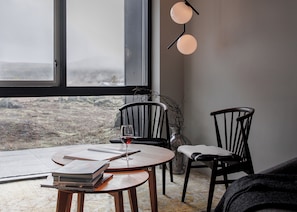 A cozy room with a large window, two chairs, a table with a wine glass, and a vase with branches.