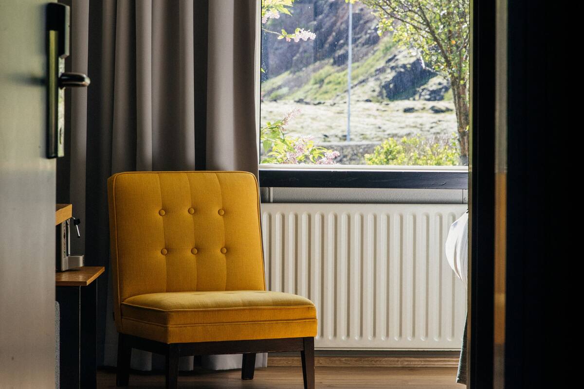 Una habitación con un sillón amarillo, un ventanal con vistas a un paisaje montañoso y un radiador.