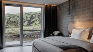 A bedroom with a large bed, a nightstand, a lamp, and a view of a mountainous landscape through a sliding door. - ION Adventure Hotel, Nesjavellir, a Member of Design Hotels (Selfoss)