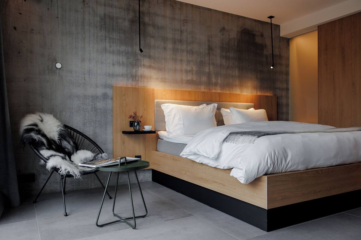Un dormitorio moderno con cabecera de madera, una cama grande, una mesita de noche con una taza y una silla con un plaid de piel.