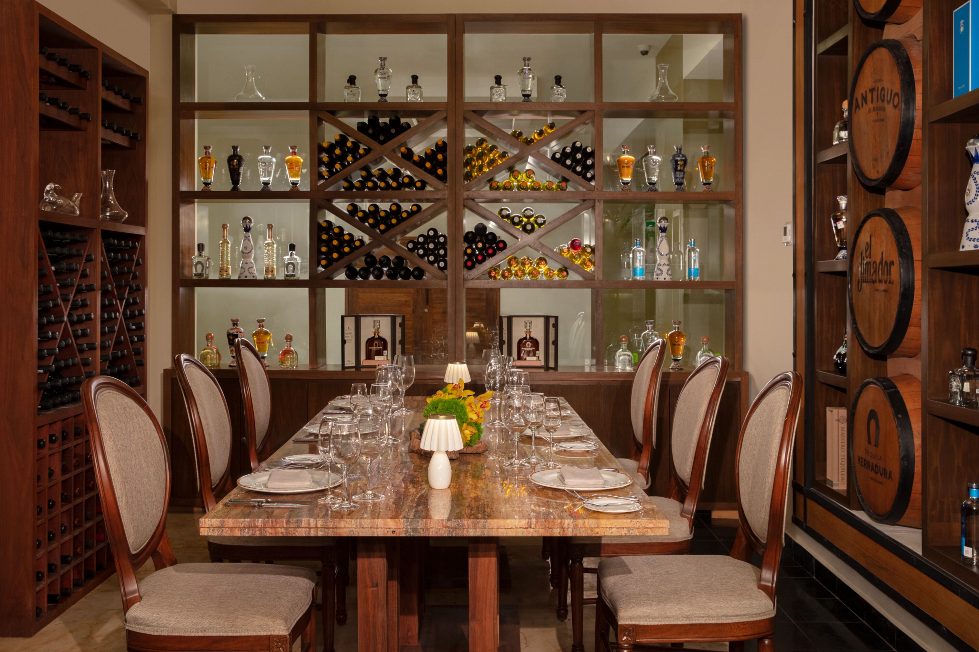 A dining area with a long table set for six, surrounded by wooden chairs. Behind the table, there are shelves with wine bottles and glassware.