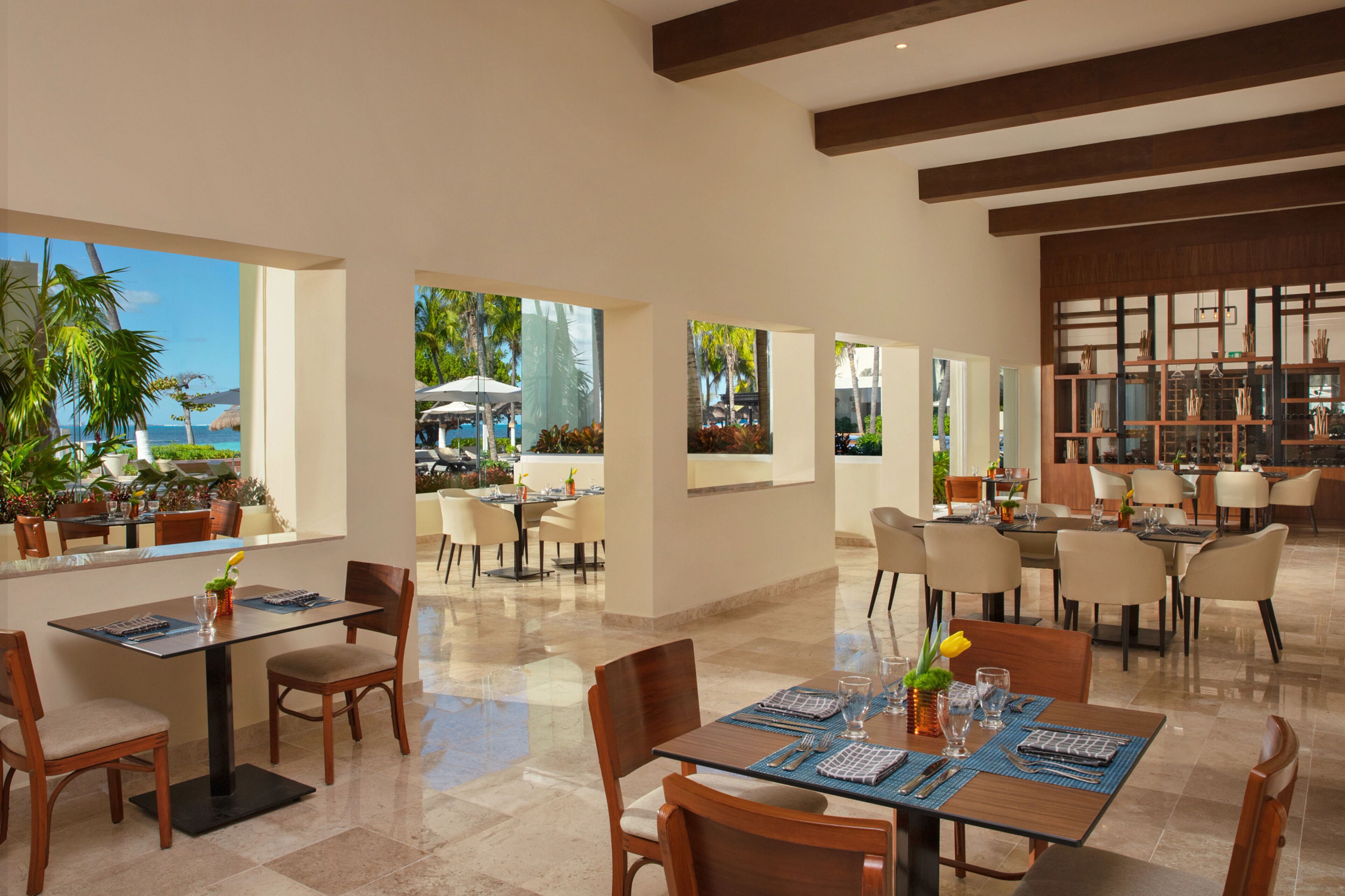 A modern dining area with tables and chairs, large windows offering views of the ocean and palm trees, and a bar area with shelves.