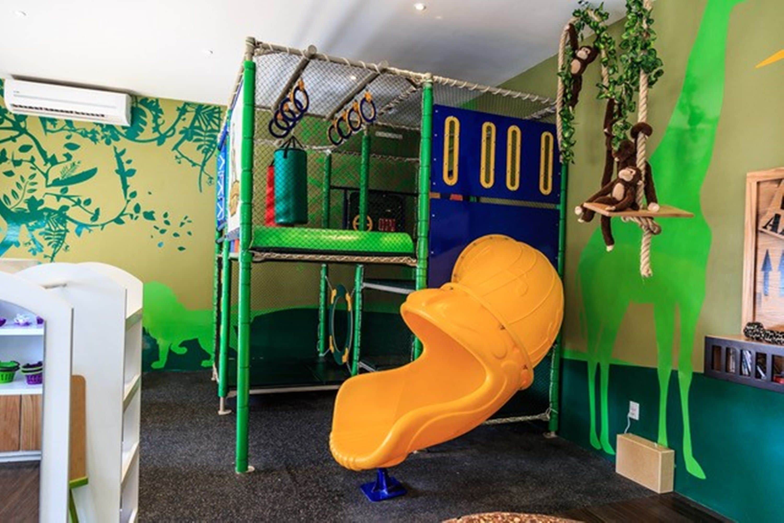 Espaço infantil com escorrega, rede de escalada e decoração com tema de macaco.