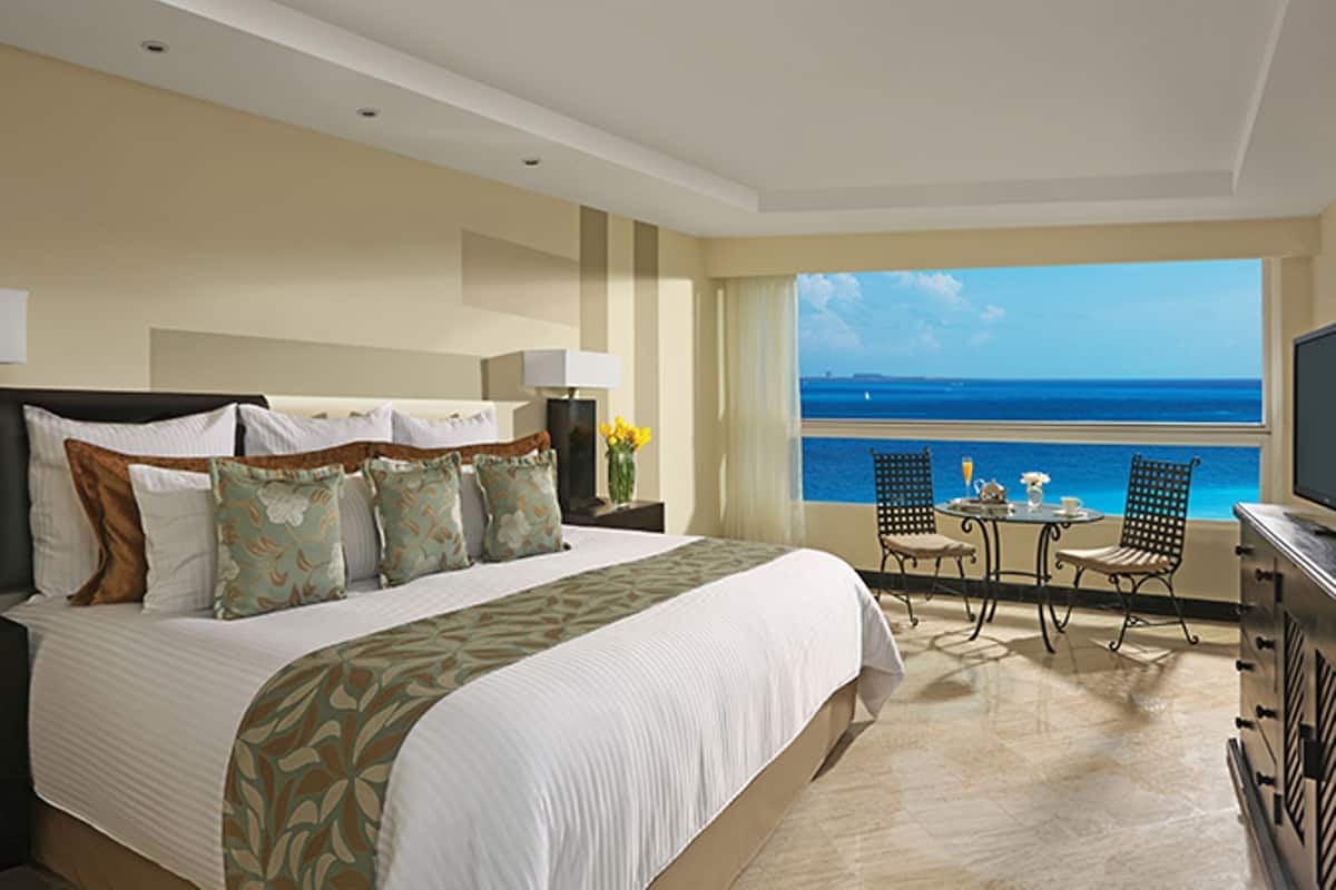 A hotel room with a large bed, a TV, a balcony with a view of the sea, and a small table with chairs.
