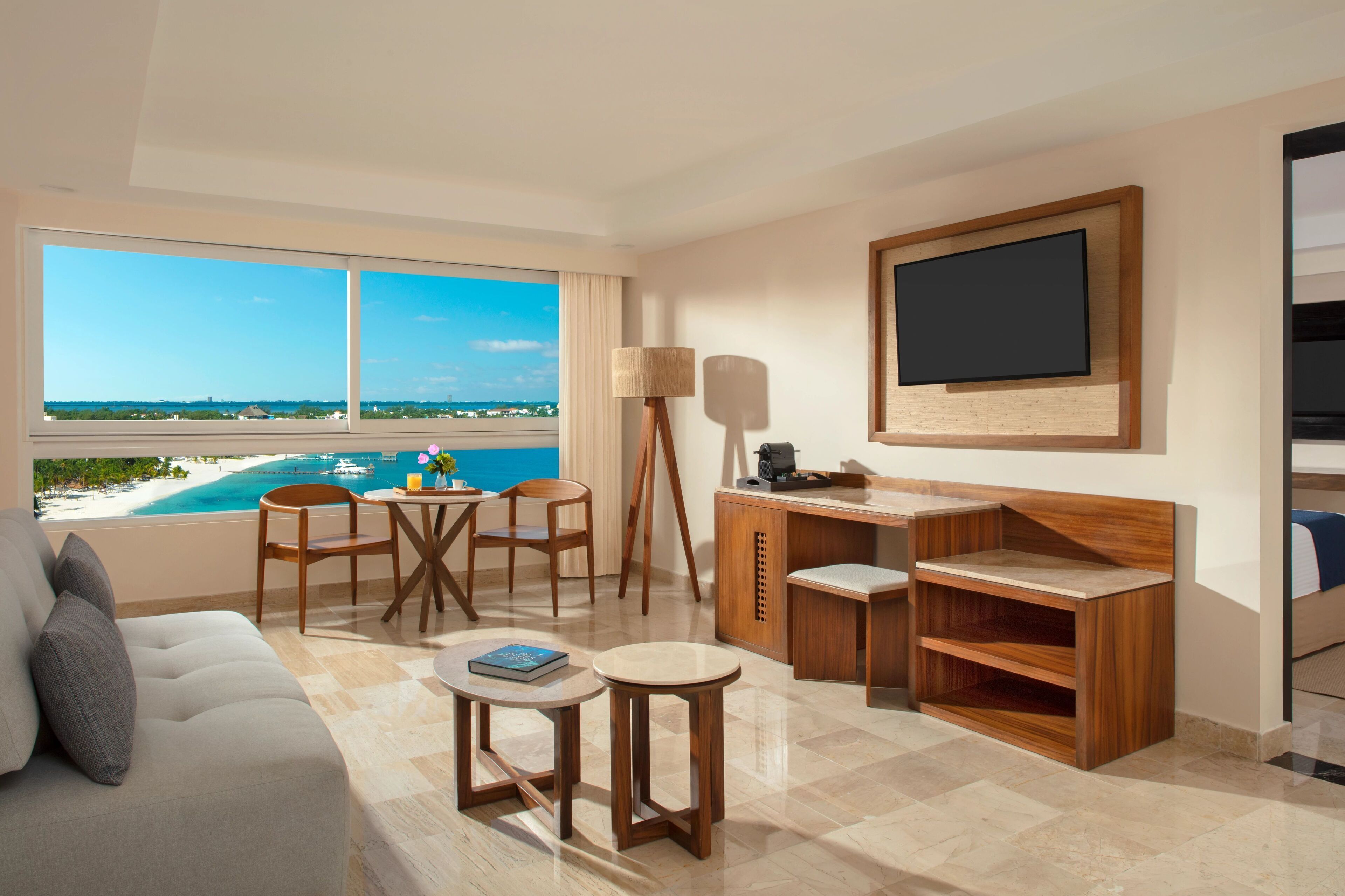 A modern hotel room with a large window offering a view of the beach and ocean. There is a wooden desk, a comfortable sofa, and a small round table.