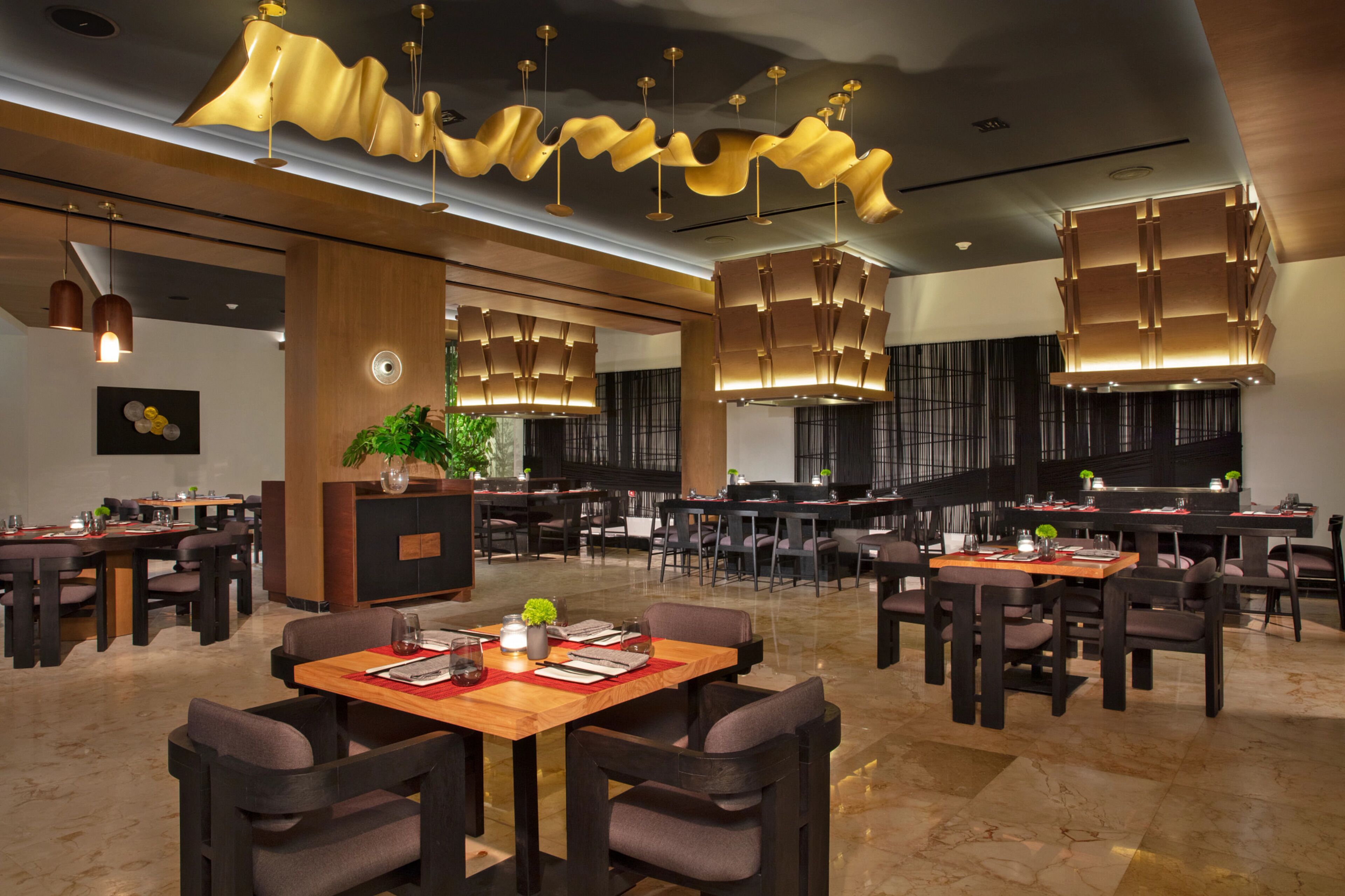 A modern restaurant with multiple dining tables and chairs, ambient lighting, and a central plant.