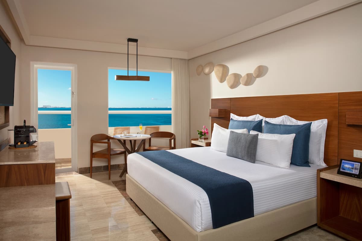 A hotel room with a large bed, a desk with a coffee machine, a chair, and a view of the sea.