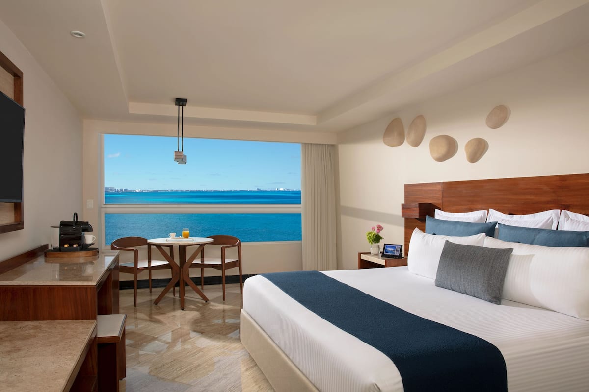 A hotel room with a large bed, a desk with a coffee maker, a chair, a small table with a cup and orange juice, and a view of the ocean.