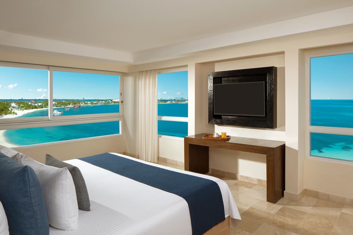 A hotel room with a bed, a TV, a small table, and large windows offering a view of the ocean and a distant cityscape.