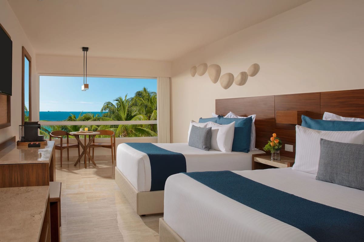 A hotel room with two beds, a desk, and a chair. There is a view of the ocean and palm trees through large windows.