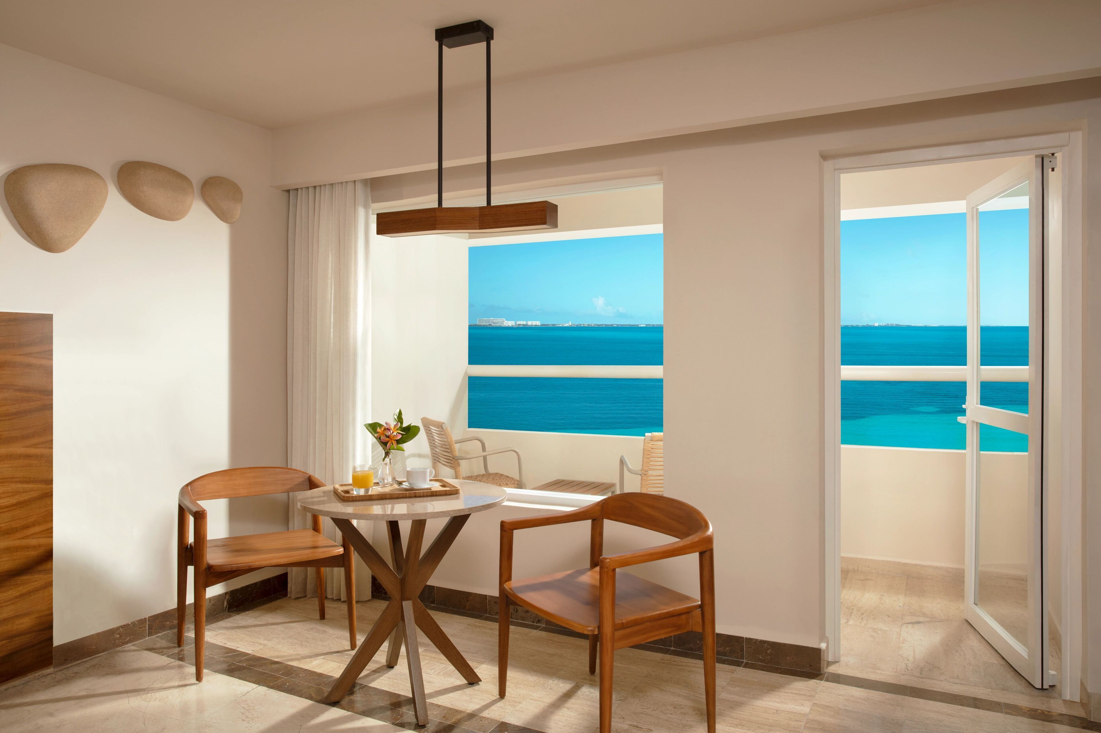 A room with a balcony overlooking a pool and the sea, featuring a round table with chairs, a hanging light fixture, and a view of the ocean.