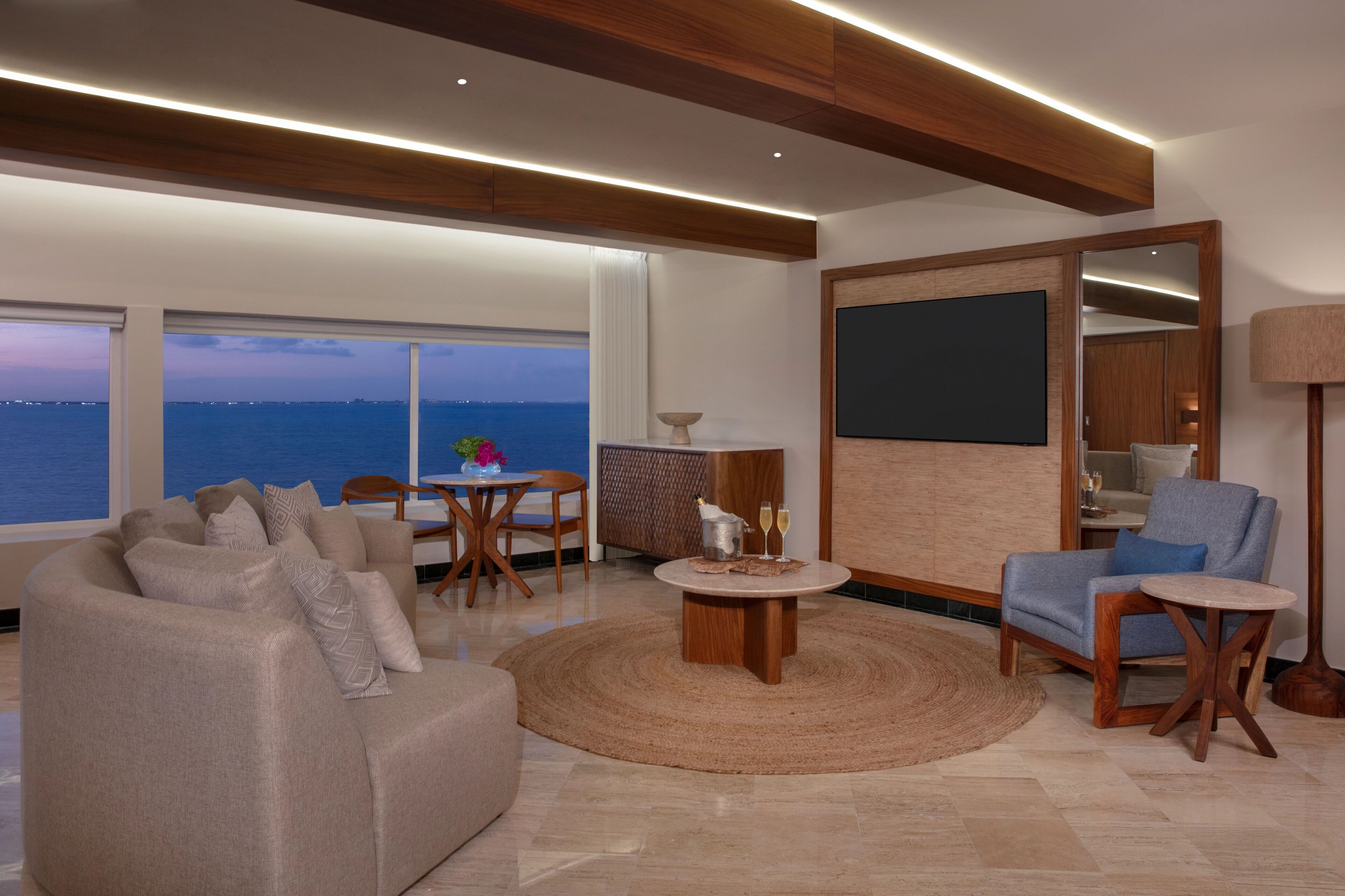 Una sala moderna con sofá, sillones, una mesa de centro y un comedor. Hay un ventanal con vistas al mar.