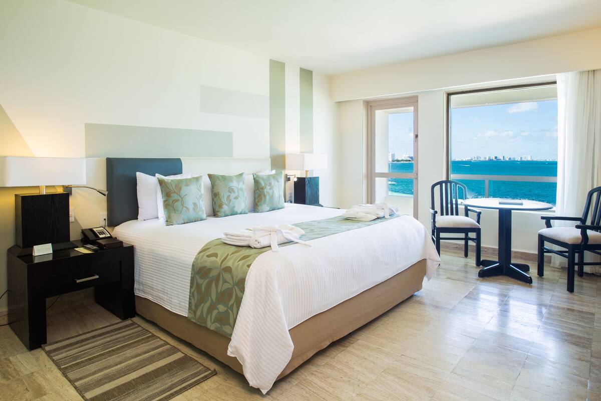 A hotel room with a large bed, a desk with a lamp, a chair, a small table, and a view of the sea.