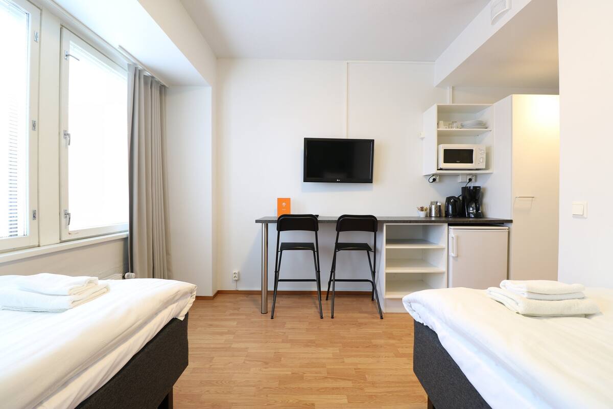 Habitación de hotel con dos camas, un escritorio con sillas, un televisor, microondas y una pequeña kitchenette.