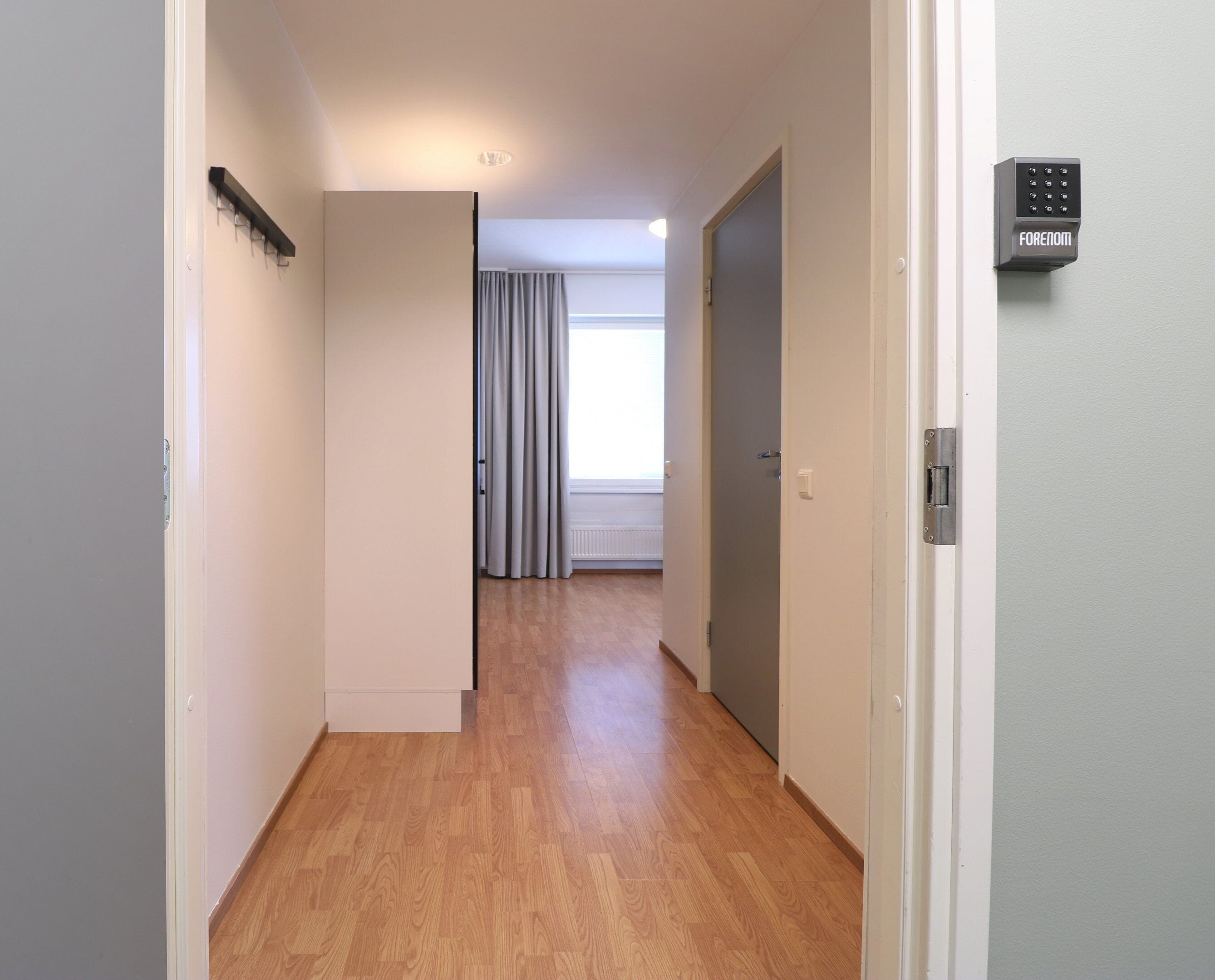A hallway with wooden flooring, a door with a security keypad, and a window with curtains.