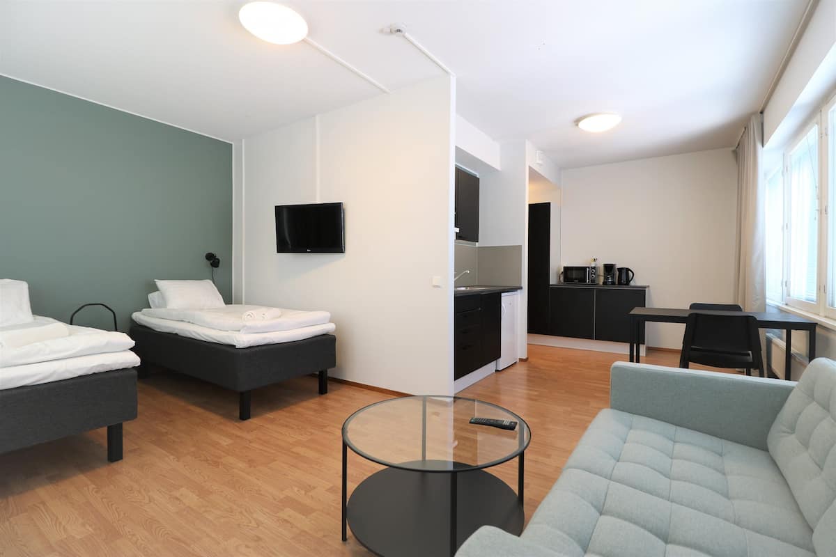Habitación moderna y bien iluminada con sofá, dos camas, una mesita de vidrio, televisión y una kitchenette al fondo.