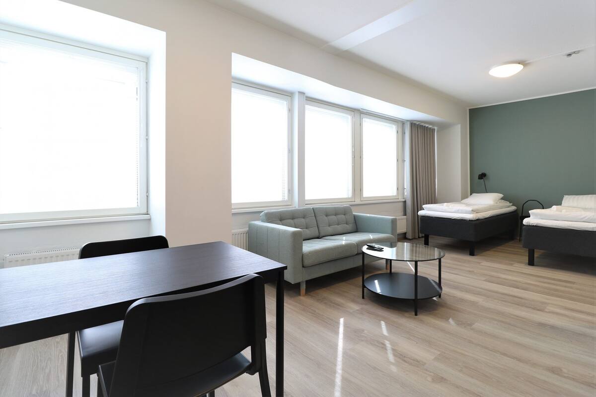 Una sala de estar moderna y bien iluminada con un sofá, dos camas, una mesa de comedor y una mesa de centro.