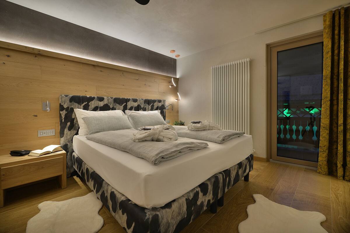 A modern bedroom with a wooden headboard, a large bed with white linens, a nightstand with a book and remote, and a view of a cityscape through a window.