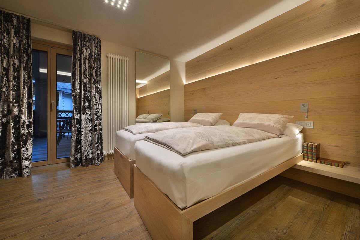 A modern bedroom with two beds, a wooden headboard, a built-in shelf, and a view through a glass door.