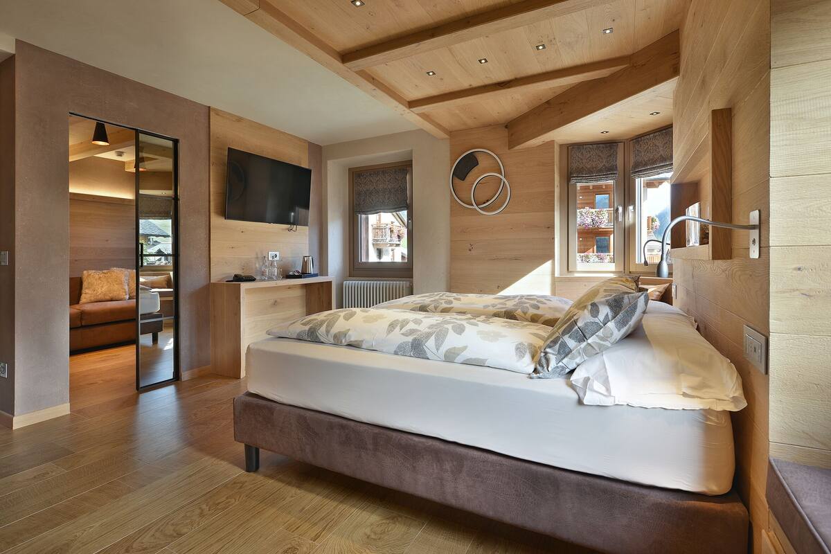 A modern bedroom with a wooden ceiling, a large bed with patterned bedding, a TV mounted on the wall, and a view of the outside through a window.