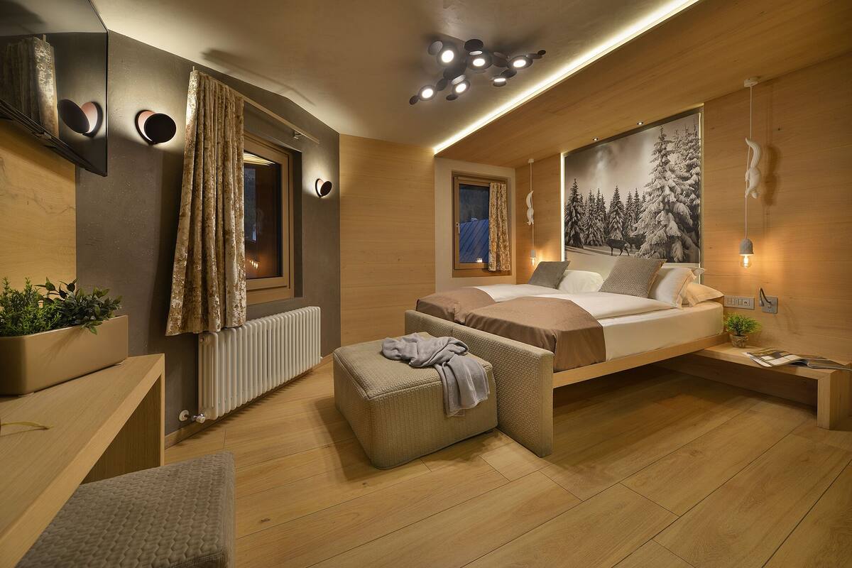 A modern bedroom with a bed, a small seating area, a window with curtains, a radiator, and a wall-mounted light fixture.
