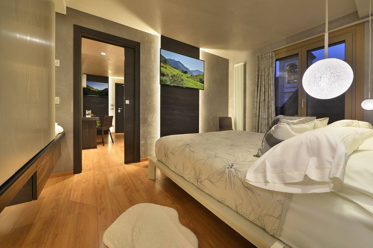 A modern bedroom with a large bed, a TV mounted on the wall, and a view of the outdoors.