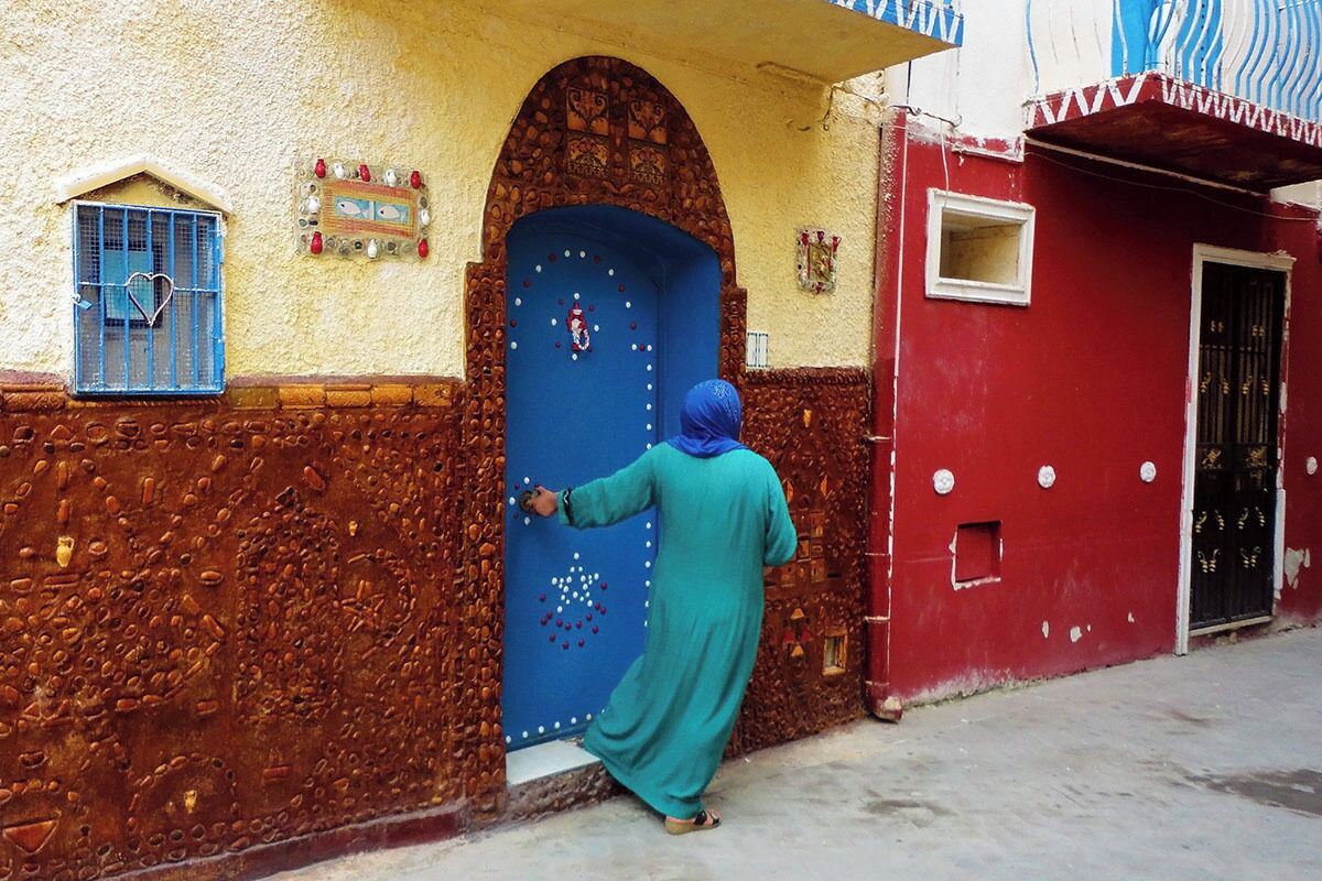 Une personne vêtue d’un costume traditionnel ouvre une porte bleue ornée d’éléments décoratifs.