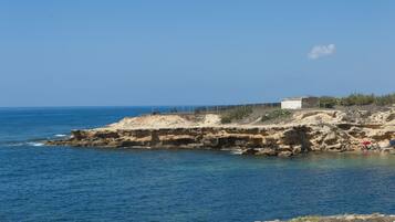 Un paysage côtier avec une rive rocheuse, une mer bleue et limpide, et un petit bâtiment perché sur la falaise.