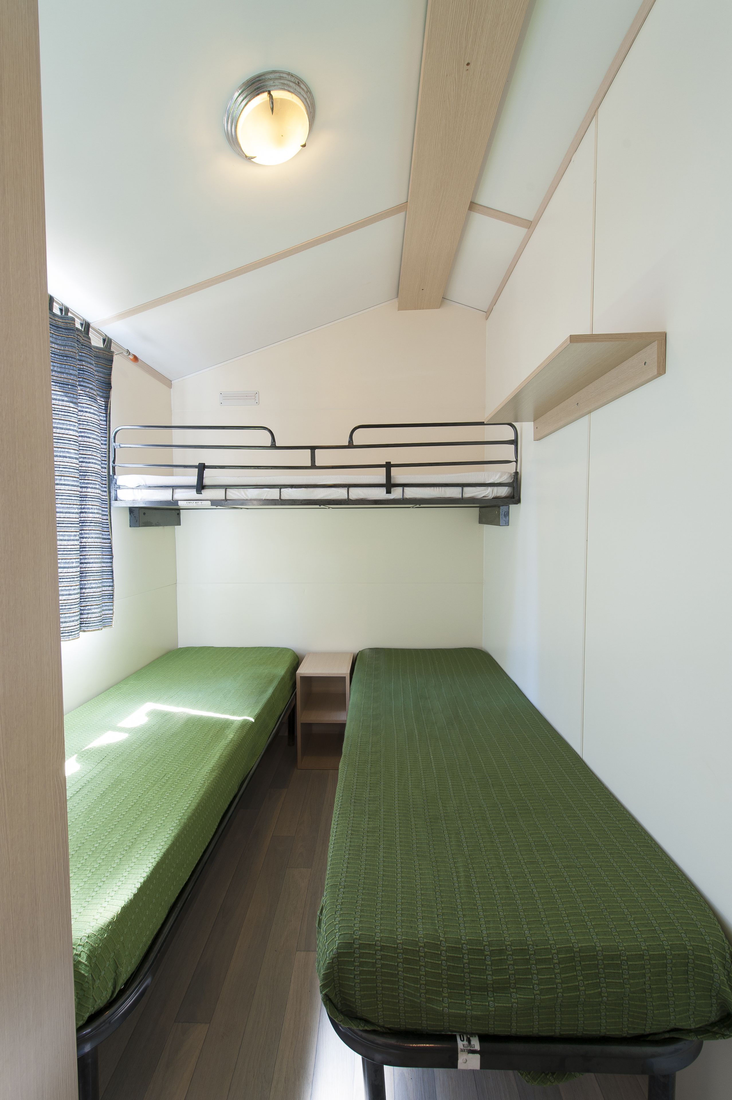 Una habitación pequeña y funcional con literas y una cama individual, una ventana con cortinas, y una lámpara en el techo.
