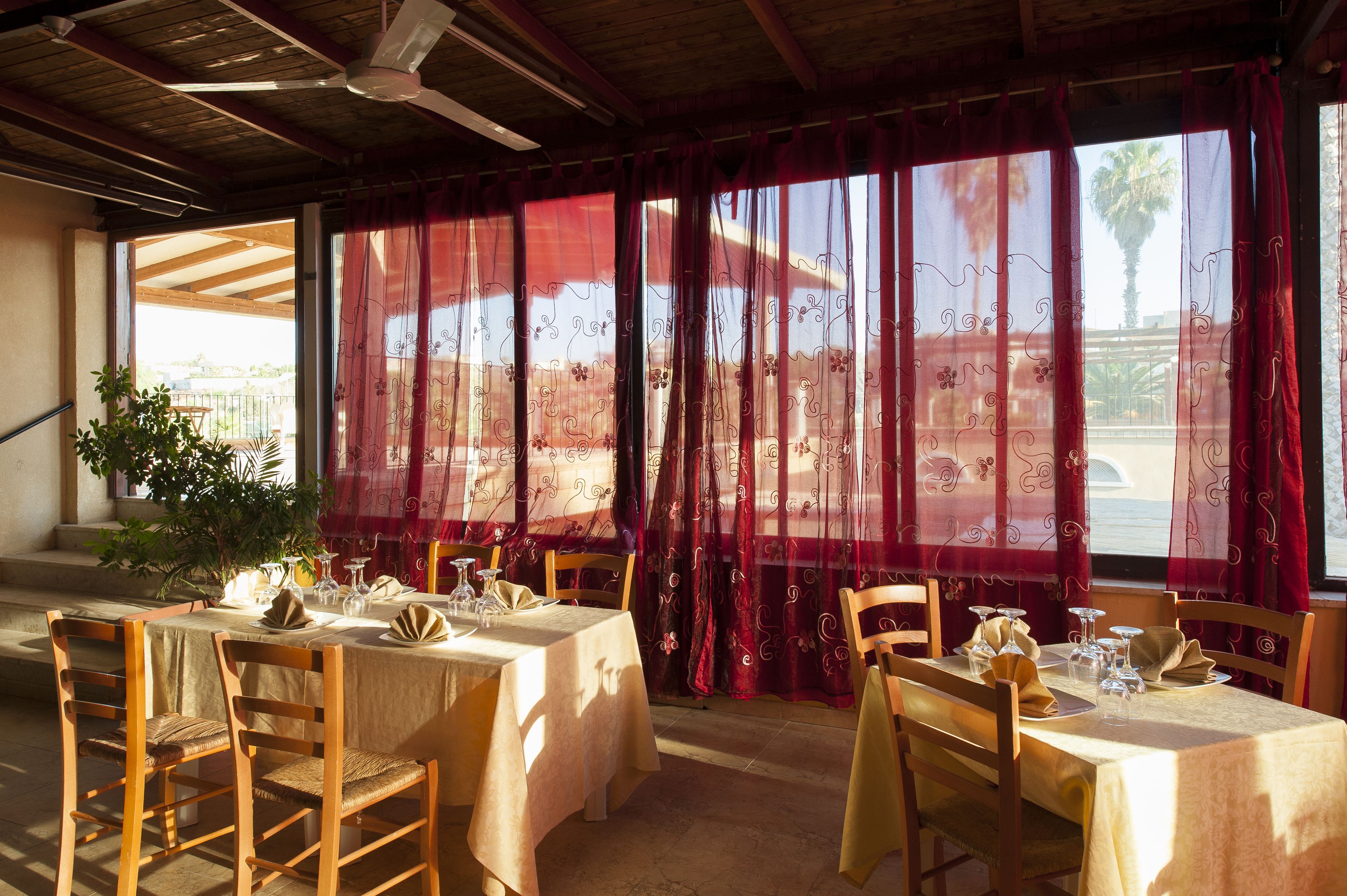 A dining area with tables set for two, wooden chairs, and a view through red curtains to the outside.