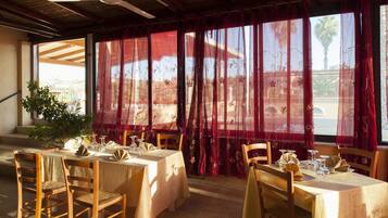 A dining area with tables set for two, wooden chairs, and a view through red curtains to the outside.