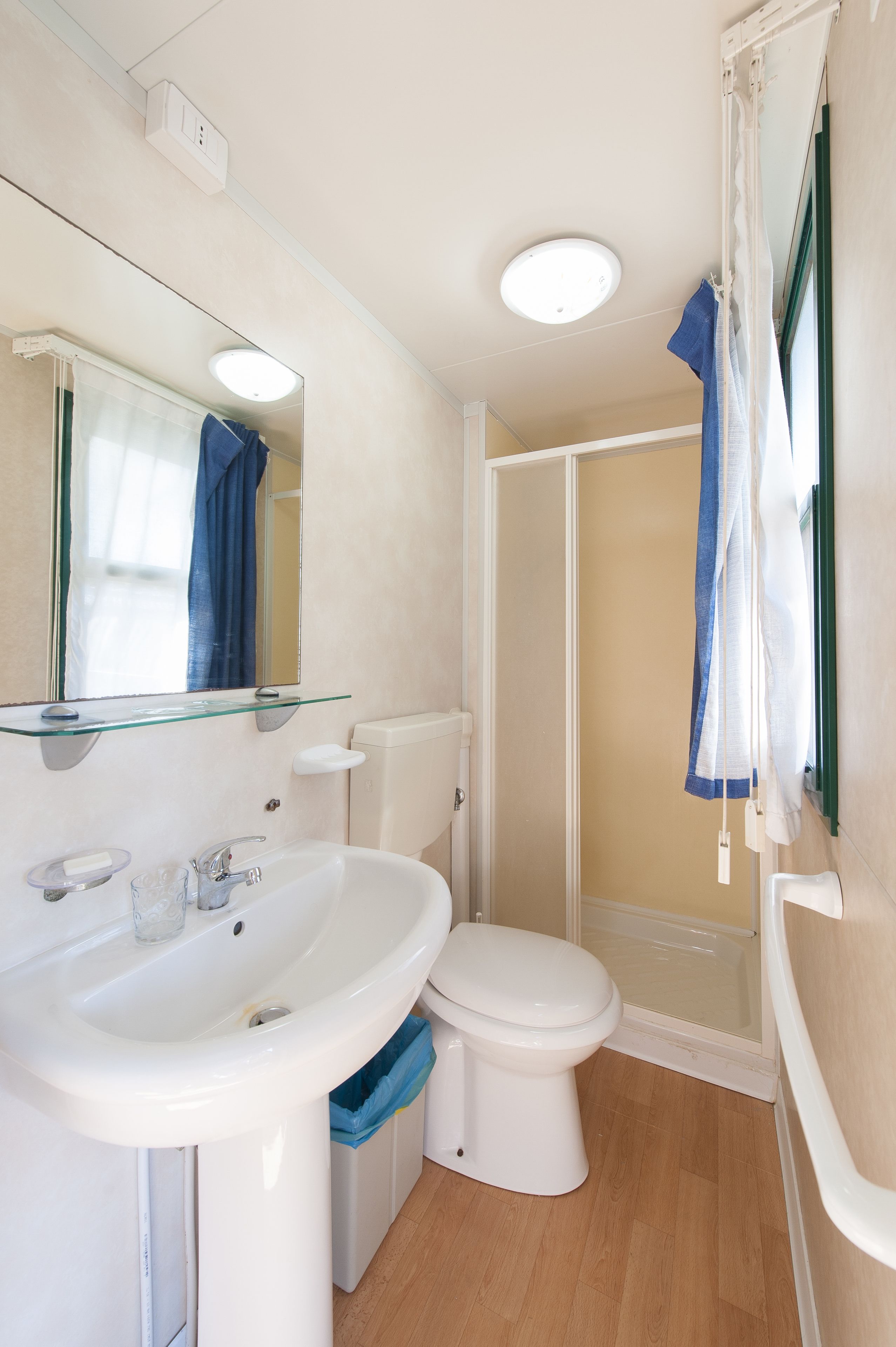 A bathroom with a white sink, toilet, and shower. There is a glass shelf above the sink and a blue towel hanging on the shower door.