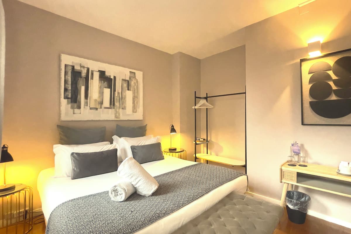 Ein modernes Hotelzimmer mit einem ordentlich bezogenen Bett, einem Nachttisch mit Lampe, einem kleinen Schreibtisch mit Wasserkocher und einem Mülleimer.