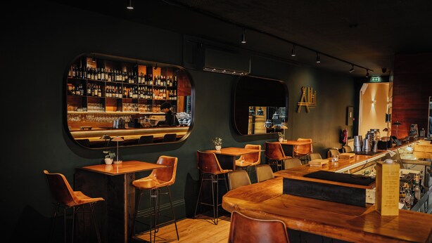Zona de bar con mesas y sillas de madera, barra y estantes llenos de botellas y vasos.
