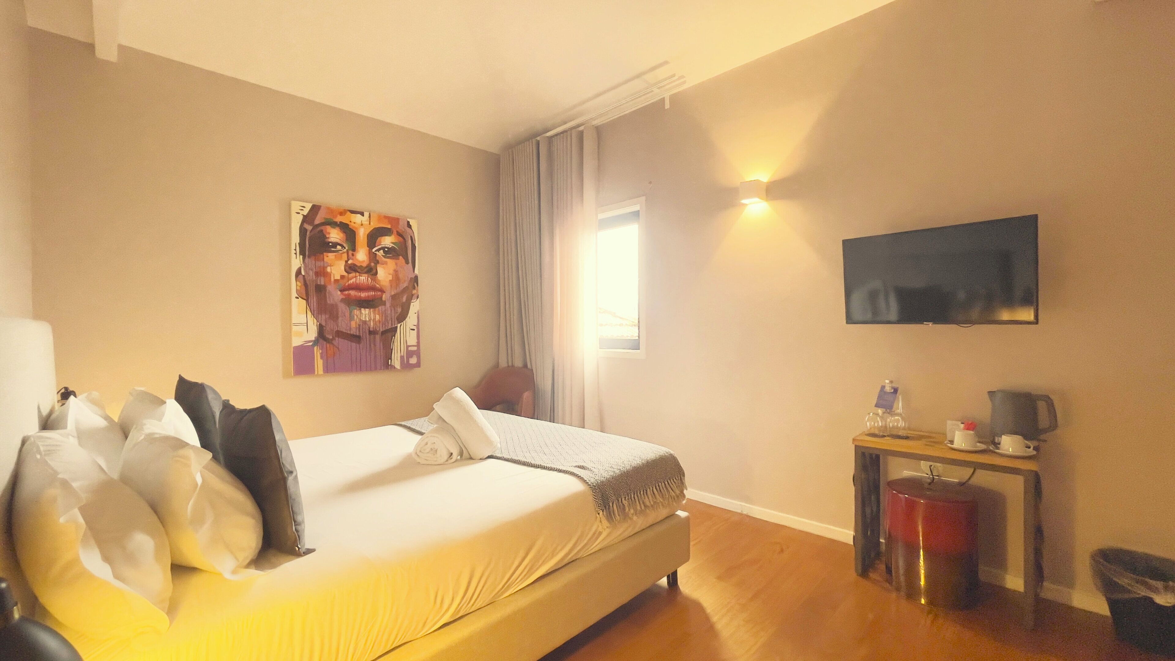 Une chambre d’hôtel avec un lit, un tableau au mur, une télévision, un bureau avec une bouilloire et une fenêtre avec des rideaux.