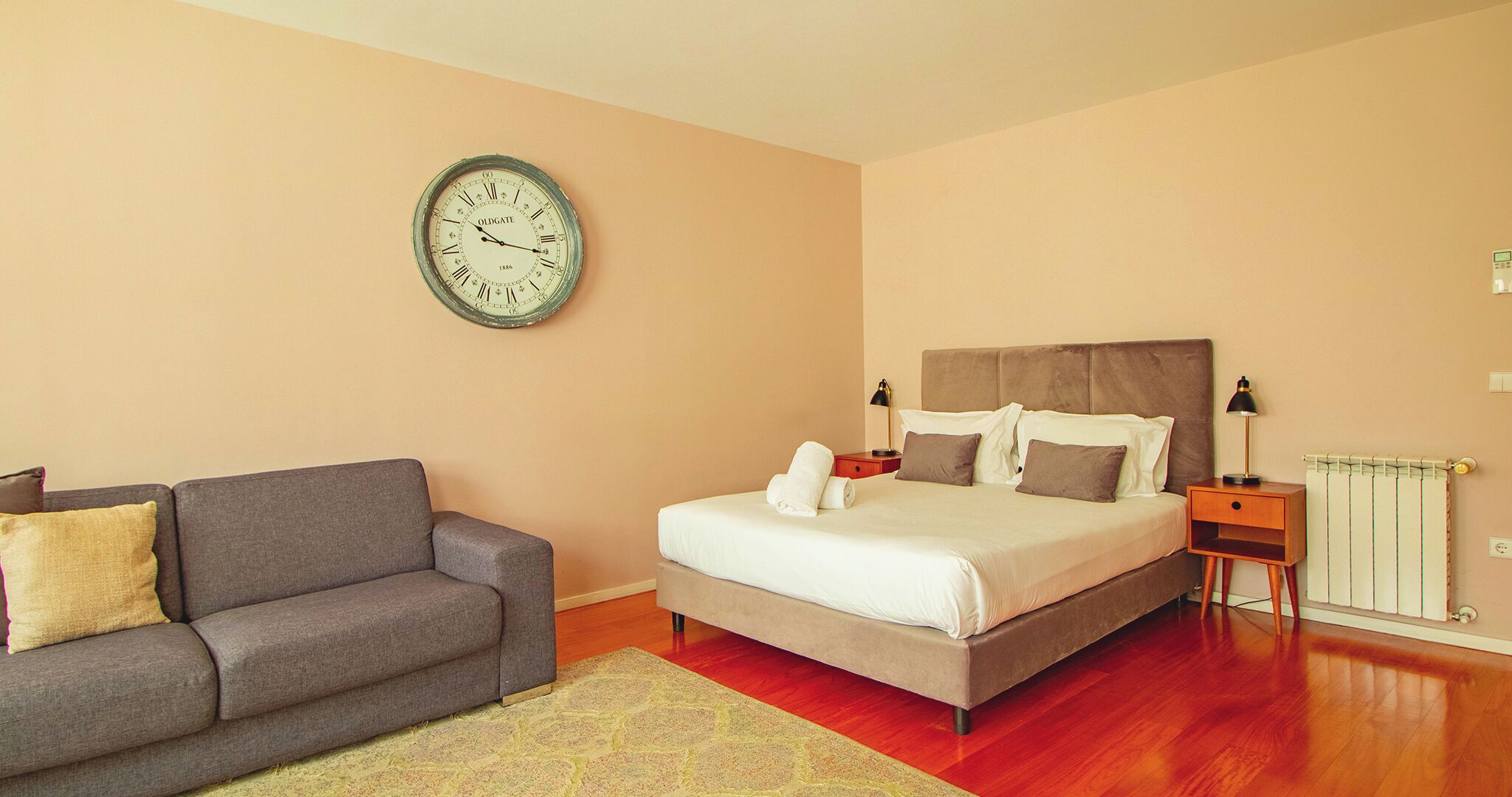 A bedroom with a bed, a grey sofa, a wall clock, and a bedside table with lamps.