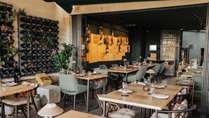 Interior de restaurante con mesas de madera, sillas acolchadas y una estantería para vinos en la pared.