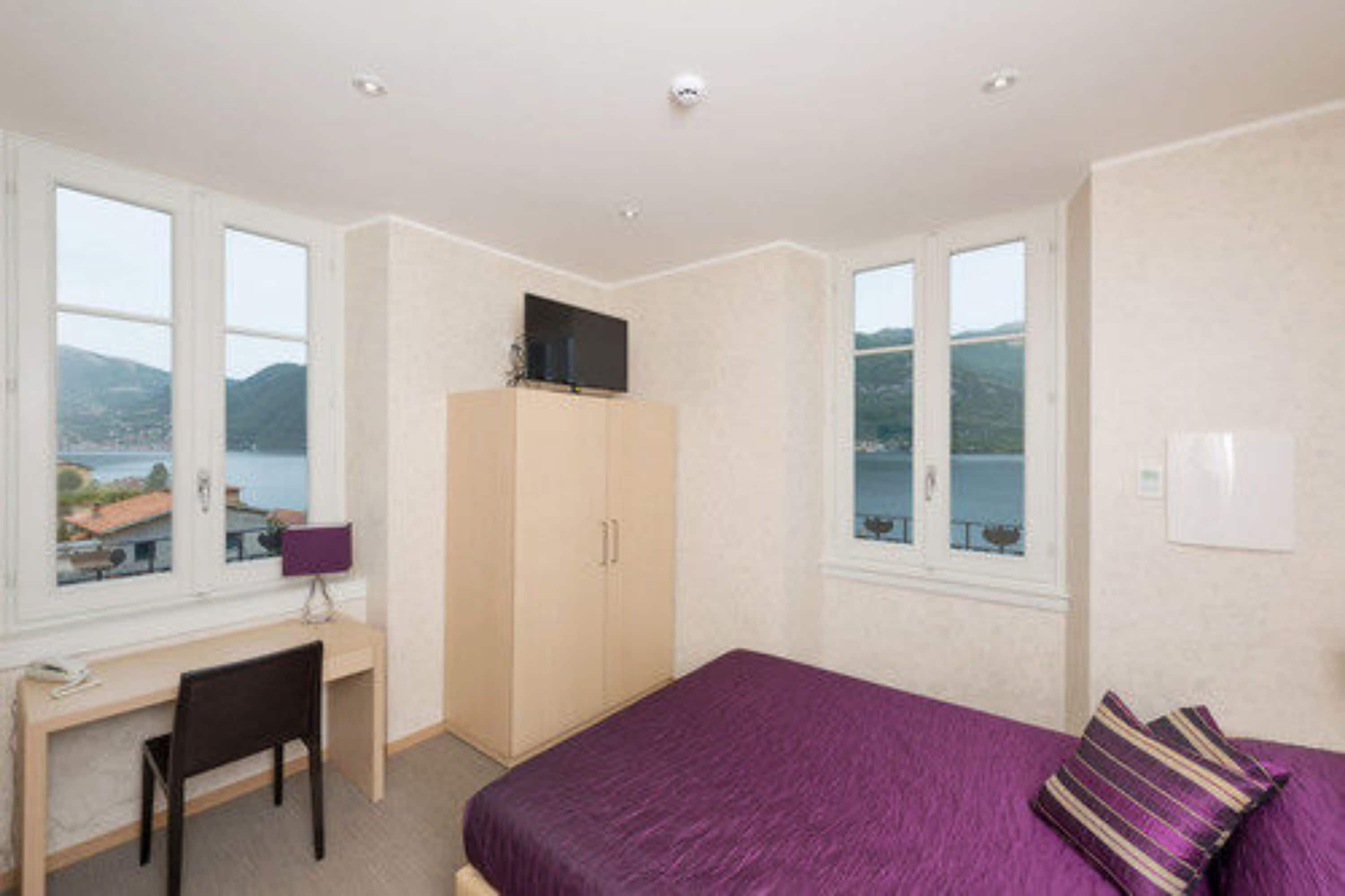 A bedroom with a bed, a desk, a chair, a wardrobe, and large windows with a view of the sea.
