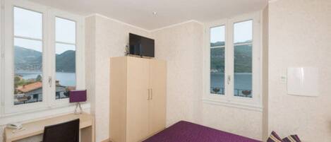 A bedroom with a bed, a desk, a chair, a wardrobe, and large windows with a view of the sea.