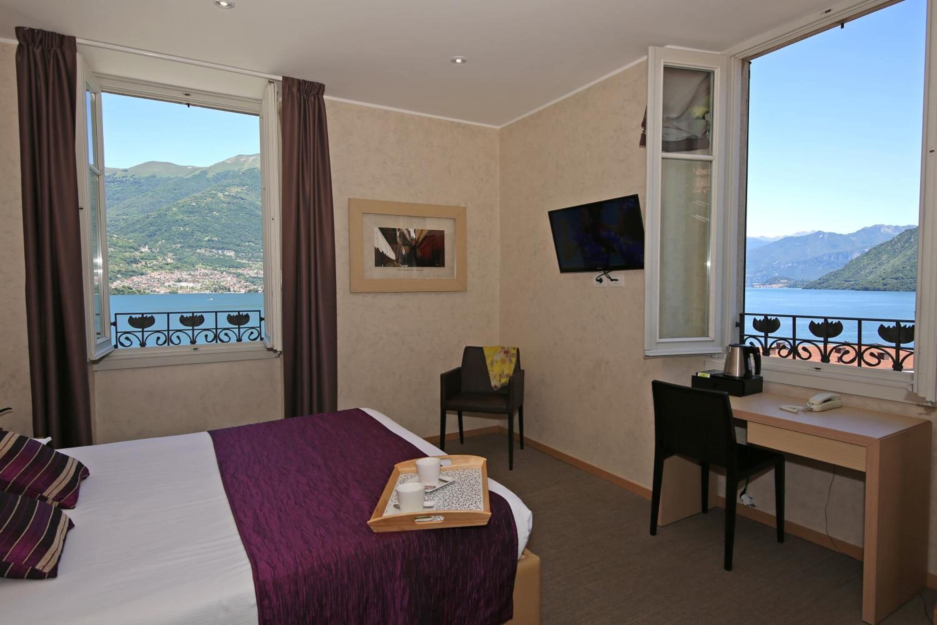 Habitación de hotel con cama, escritorio, silla, televisor y ventana con vista a montañas y un lago.