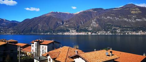 A lakeside town with terracotta-roofed buildings, a mountain in the background, and a clear blue sky.