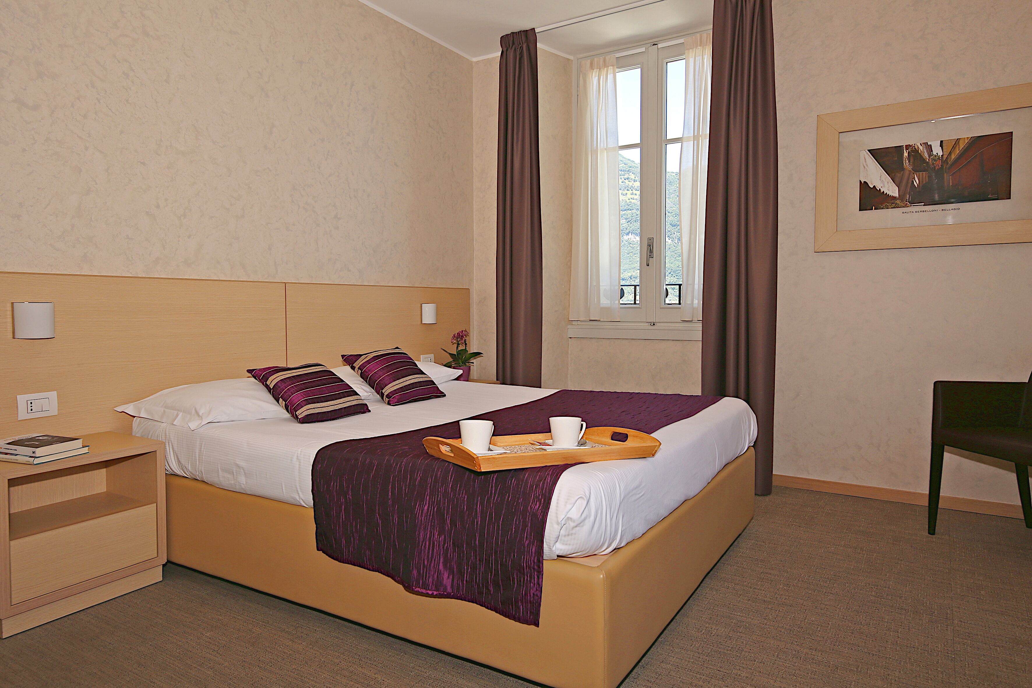 A hotel room with a bed, a nightstand, a chair, a window with curtains, and a framed picture on the wall.