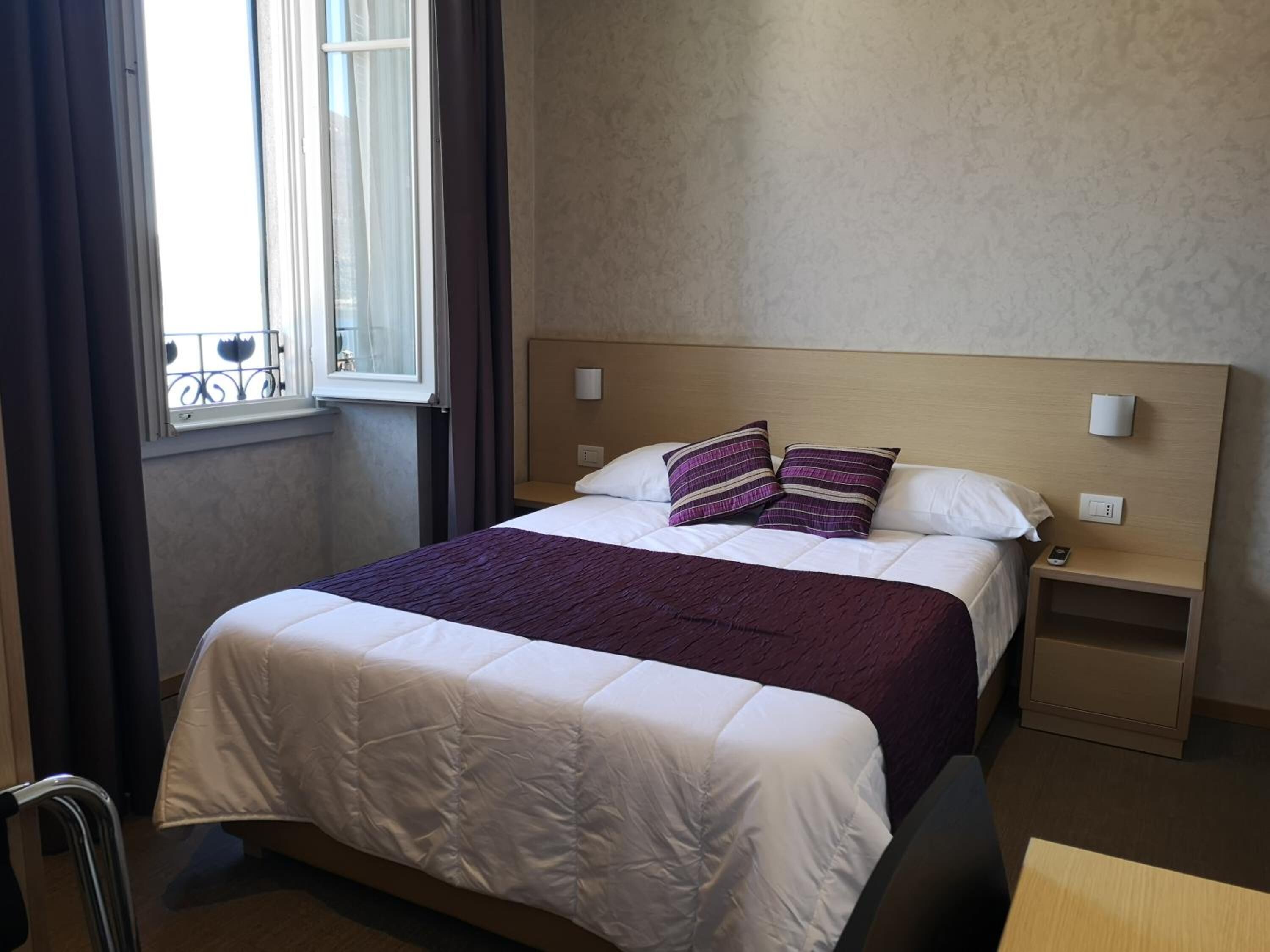 A neatly made bed with a white duvet and purple pillows, a bedside table with a lamp, and a window with curtains.