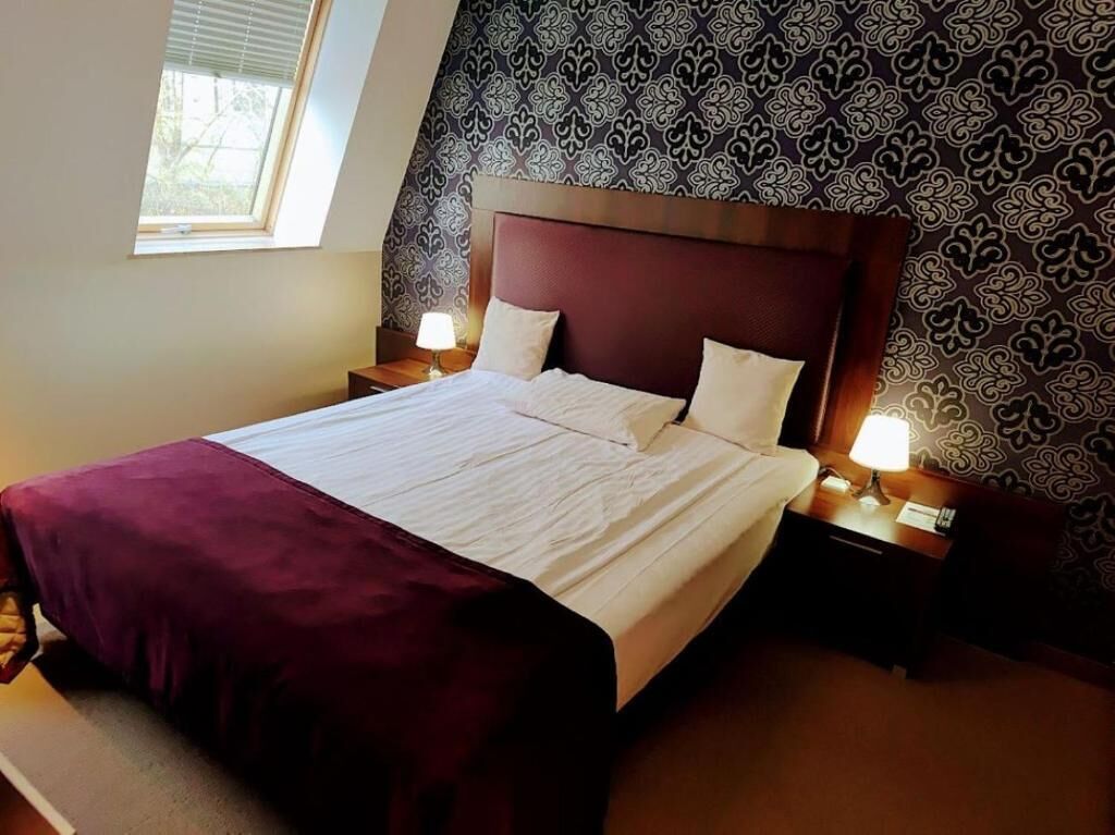 Una cama bien hecha con una colcha blanca y granate, dos almohadas y un cabecero de madera. Hay dos mesitas de noche con lámparas, una a cada lado de la cama. La habitación tiene papel tapiz estampado y una ventana con persianas.
