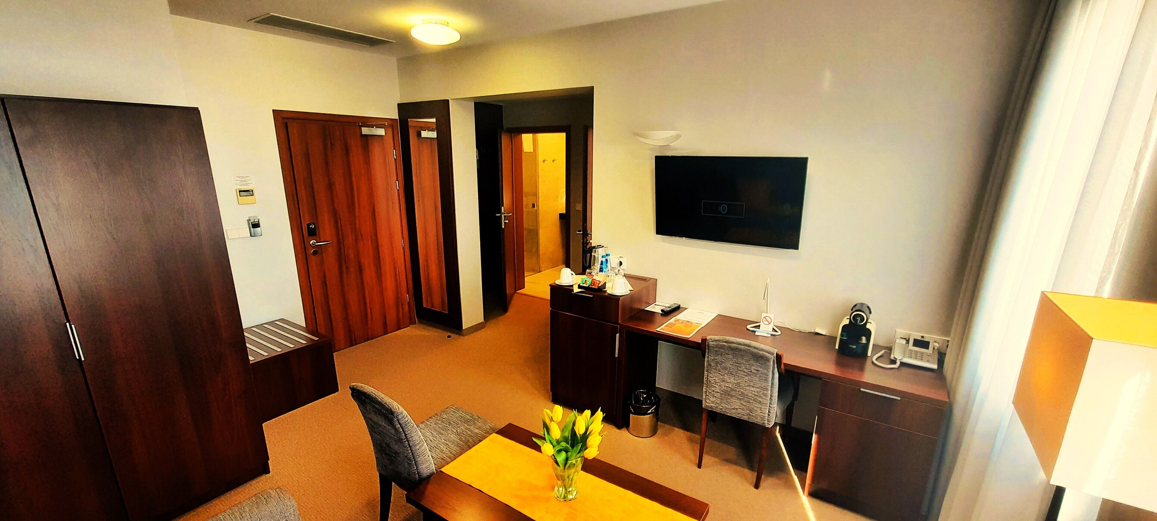 A hotel room with a wooden desk, a chair, a small table with a vase of flowers, a television mounted on the wall, and a wardrobe.