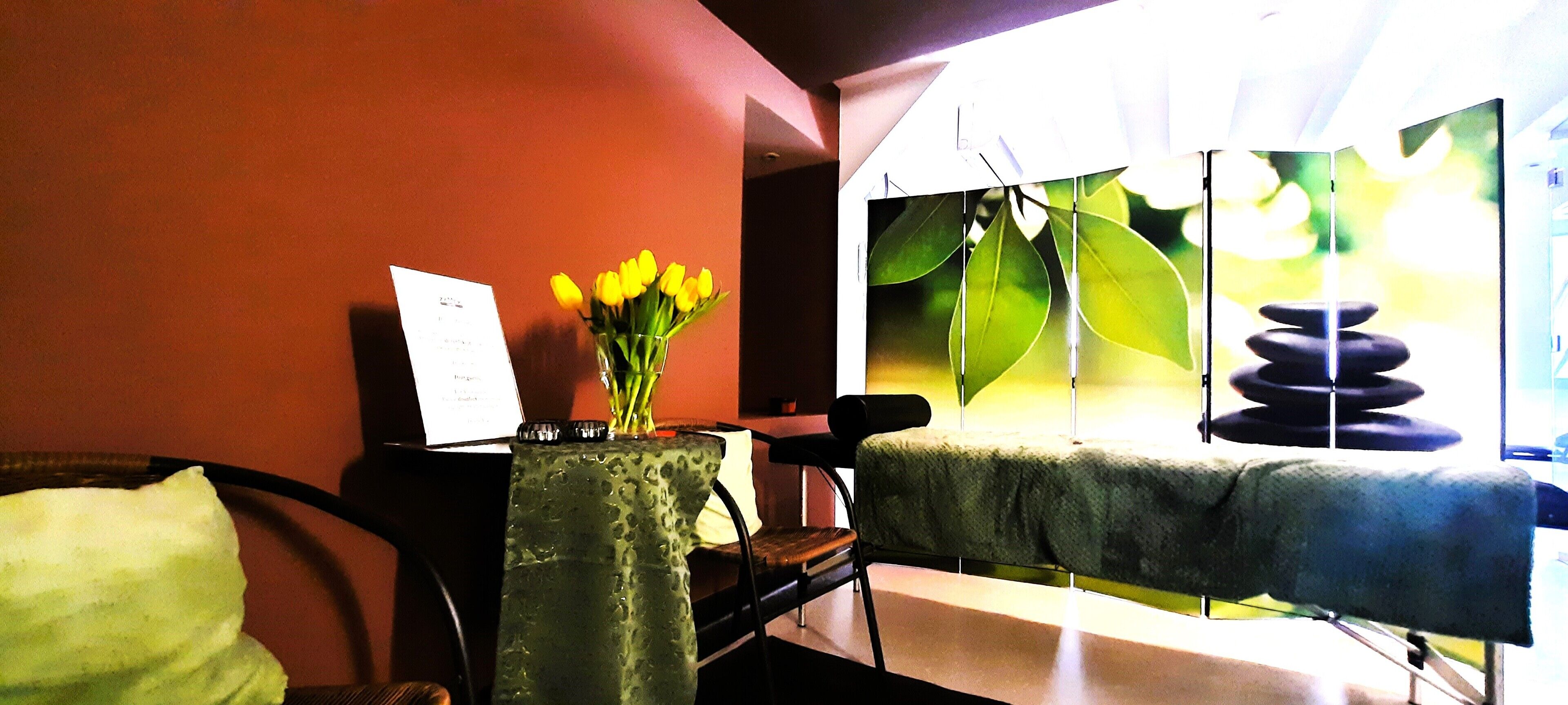 A room with a bed, a chair, a table with a vase of flowers, and a window with a view of greenery.