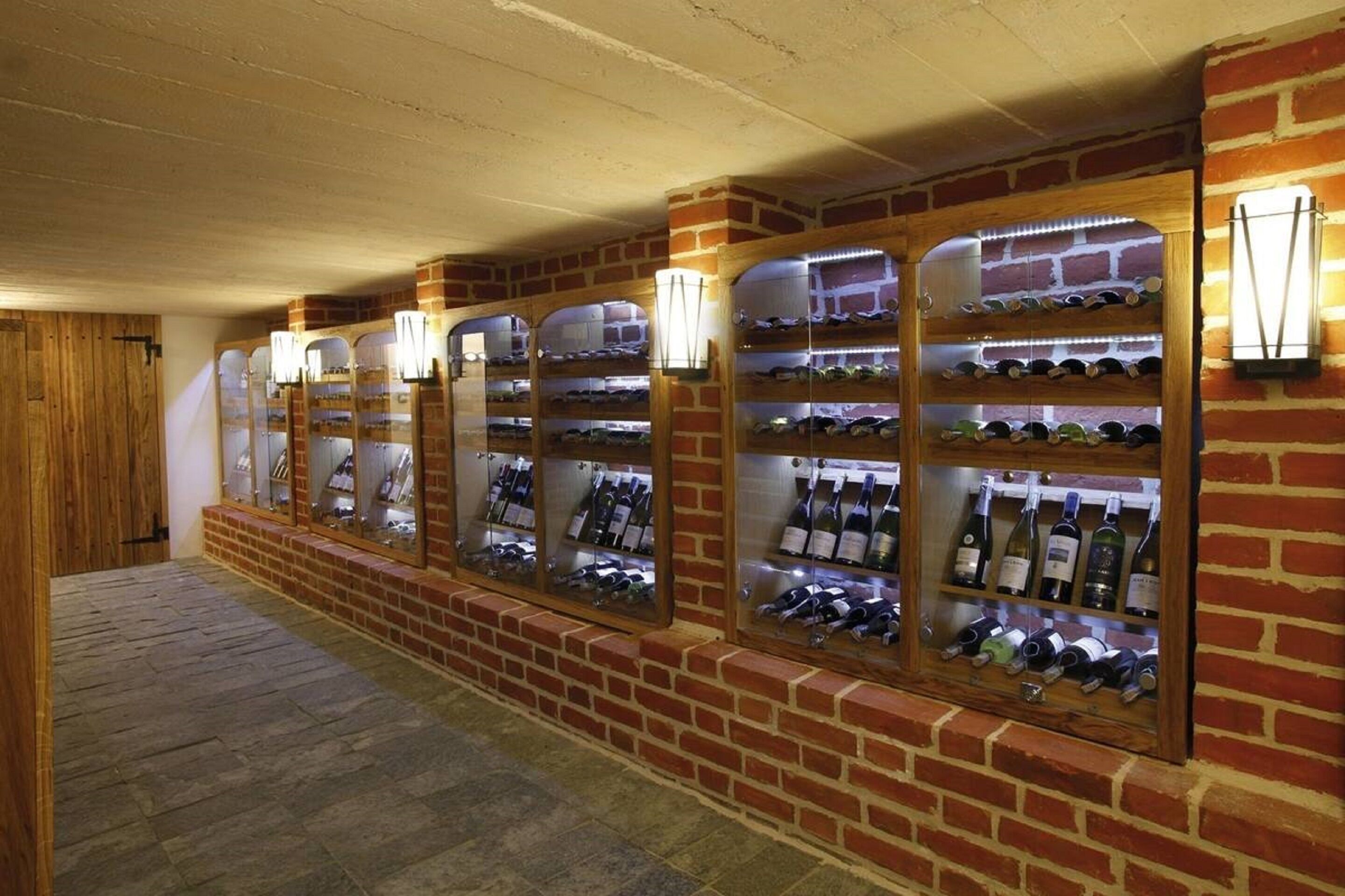 Uma adega com várias prateleiras de garrafas de vinho, iluminada por luzes fixadas na parede.