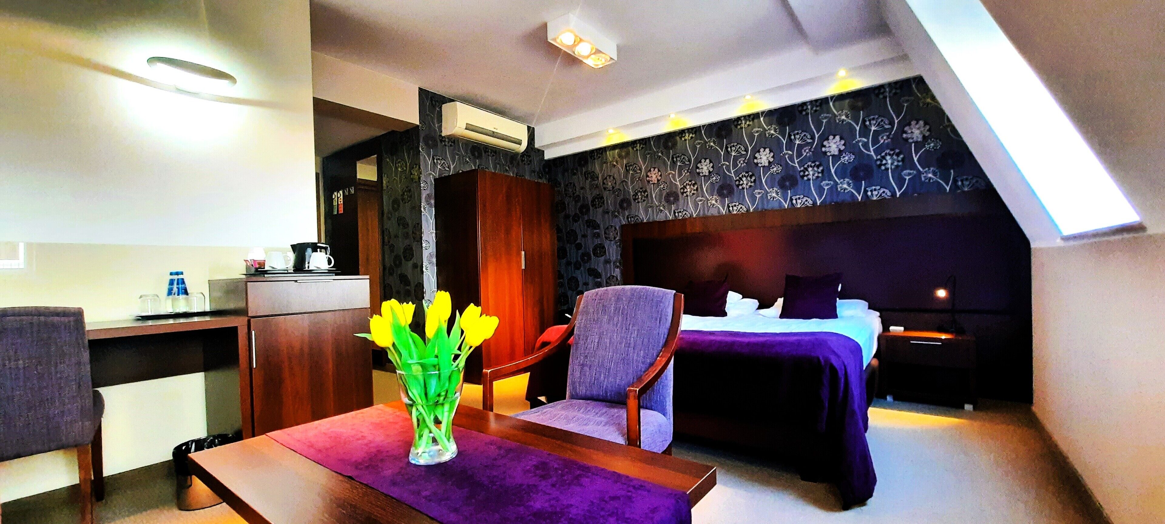 A hotel room with a bed, a desk, a chair, a small table, and a vase with flowers.