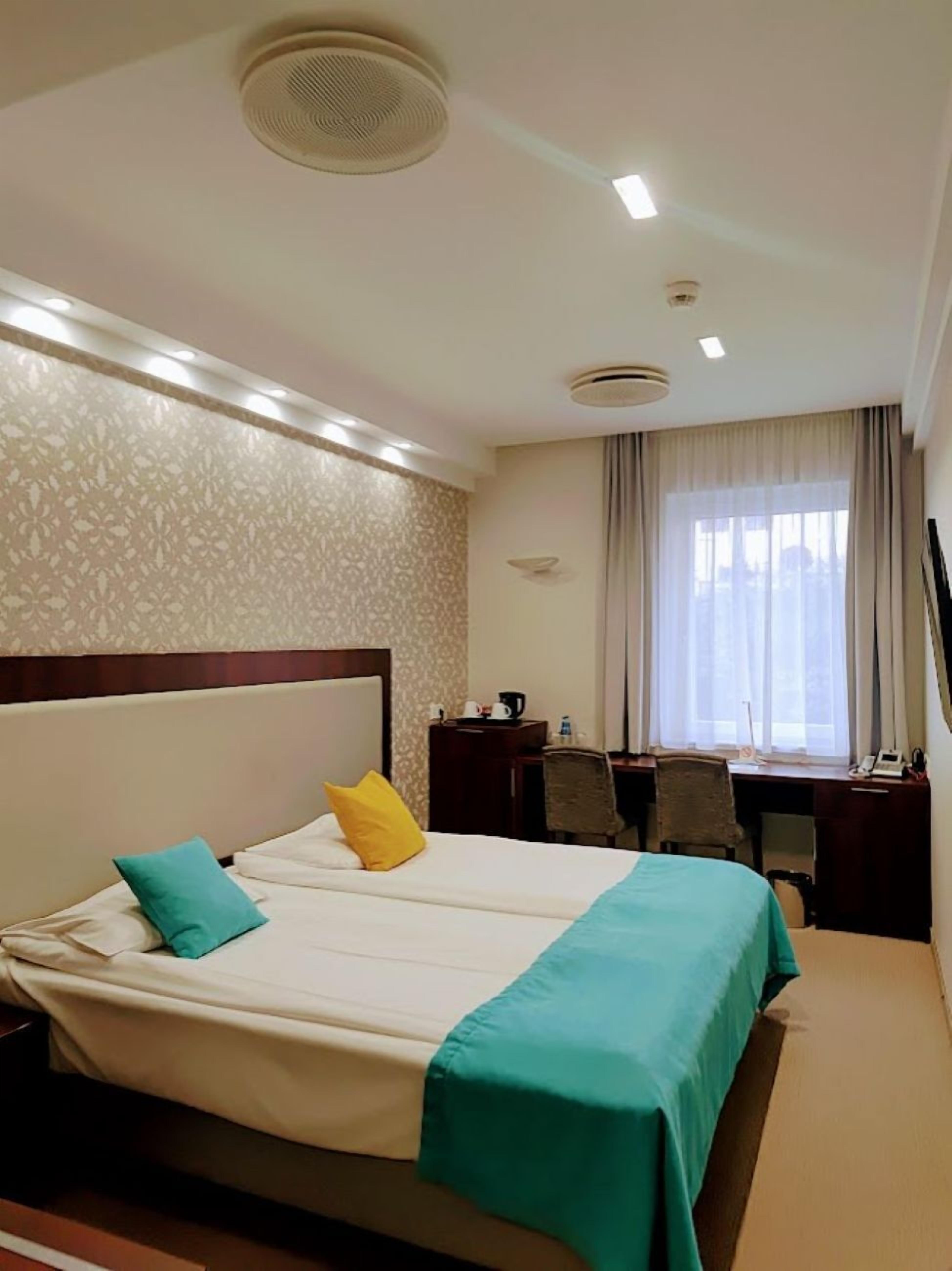 Une chambre d’hôtel avec un grand lit, un bureau et une chaise. Elle est équipée de luminaires au plafond et possède une fenêtre avec des rideaux.