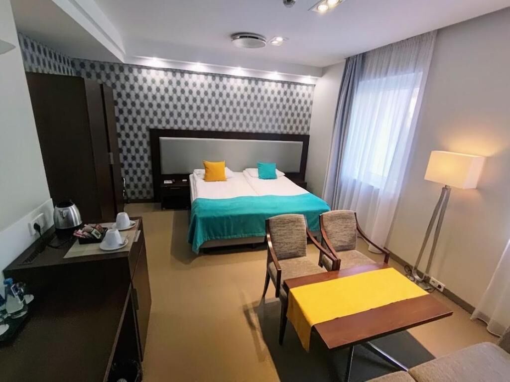 Une chambre d’hôtel comprenant un lit, deux chaises, une petite table, un bureau avec une lampe et une fenêtre avec des rideaux.